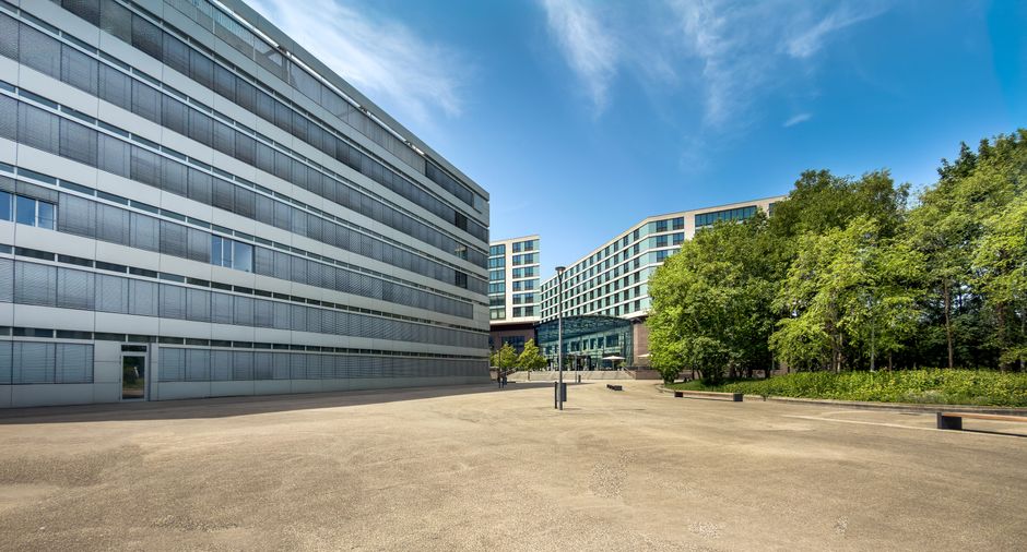 A wide view of a modern commercial office complex featuring multiple glass-fronted buildings with sleek, grey metal and glass facades. The scene is set outdoors on a sunny day with a bright blue sky a