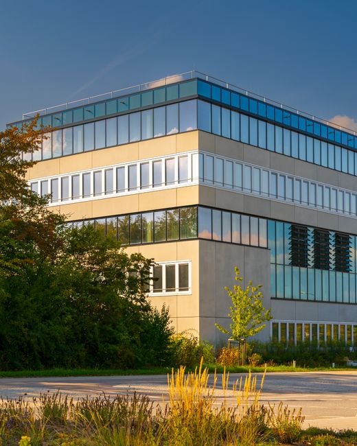 A wide view of a modern commercial office complex featuring multiple glass-fronted buildings with sleek, grey metal and glass facades. The scene is set outdoors on a sunny day with a bright blue sky a