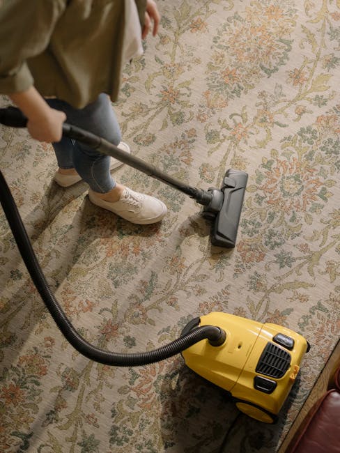 A person using their hands to place a cloth or small cleaning tool inside a yellow and black vacuum cleaner, positioned on a patterned carpet in a domestic setting. The vacuum cleaner's open top revea