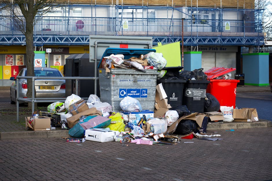 A sanitation worker from West Drayton Carpet Cleaners operating a large industrial garbage truck at dusk in an urban environment. The worker, wearing a high-visibility yellow vest with reflective stri