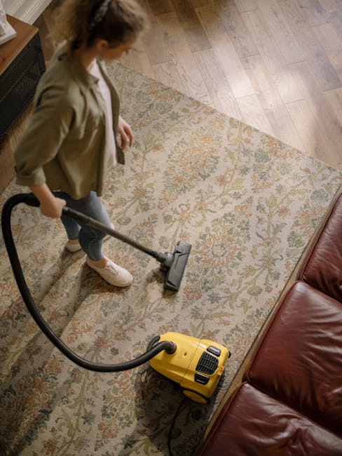 A spacious living room featuring a plush light grey carpet that appears clean and soft, with a modern grey fabric sofa against a white wall. Two small children and a man are sitting on the carpet; the