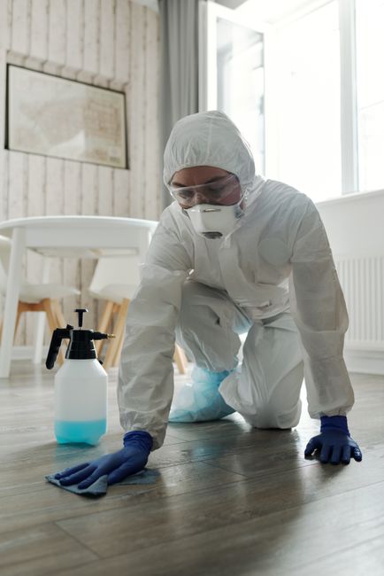 A person dressed in full protective cleaning gear, including a white coverall, gloves, a face mask, and goggles, is kneeling on a dark wooden floor in a bright, modern room with large windows. The ind